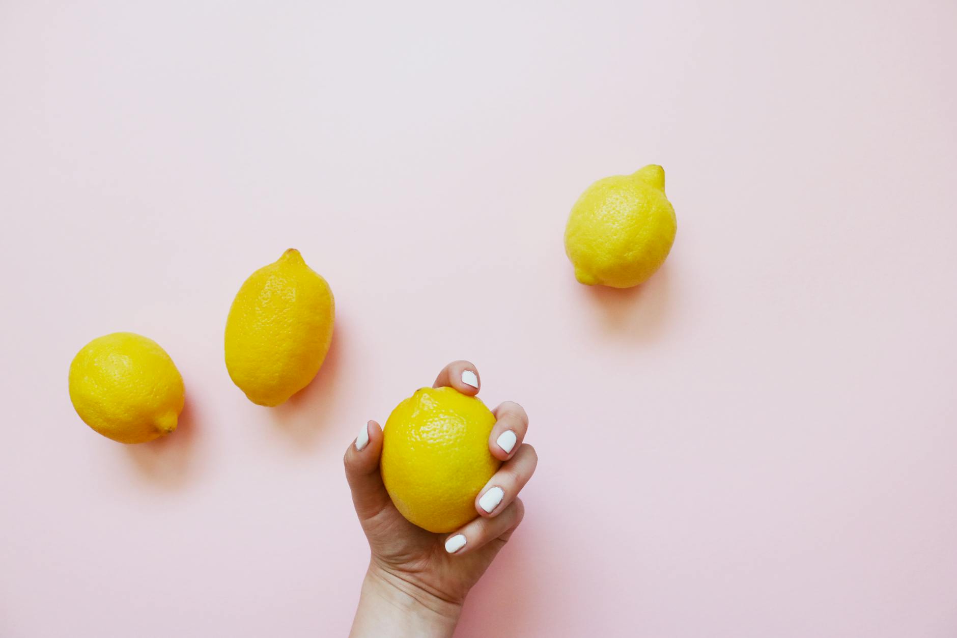 Close-up of a hand with white nails holding a lemon on a soft pink background