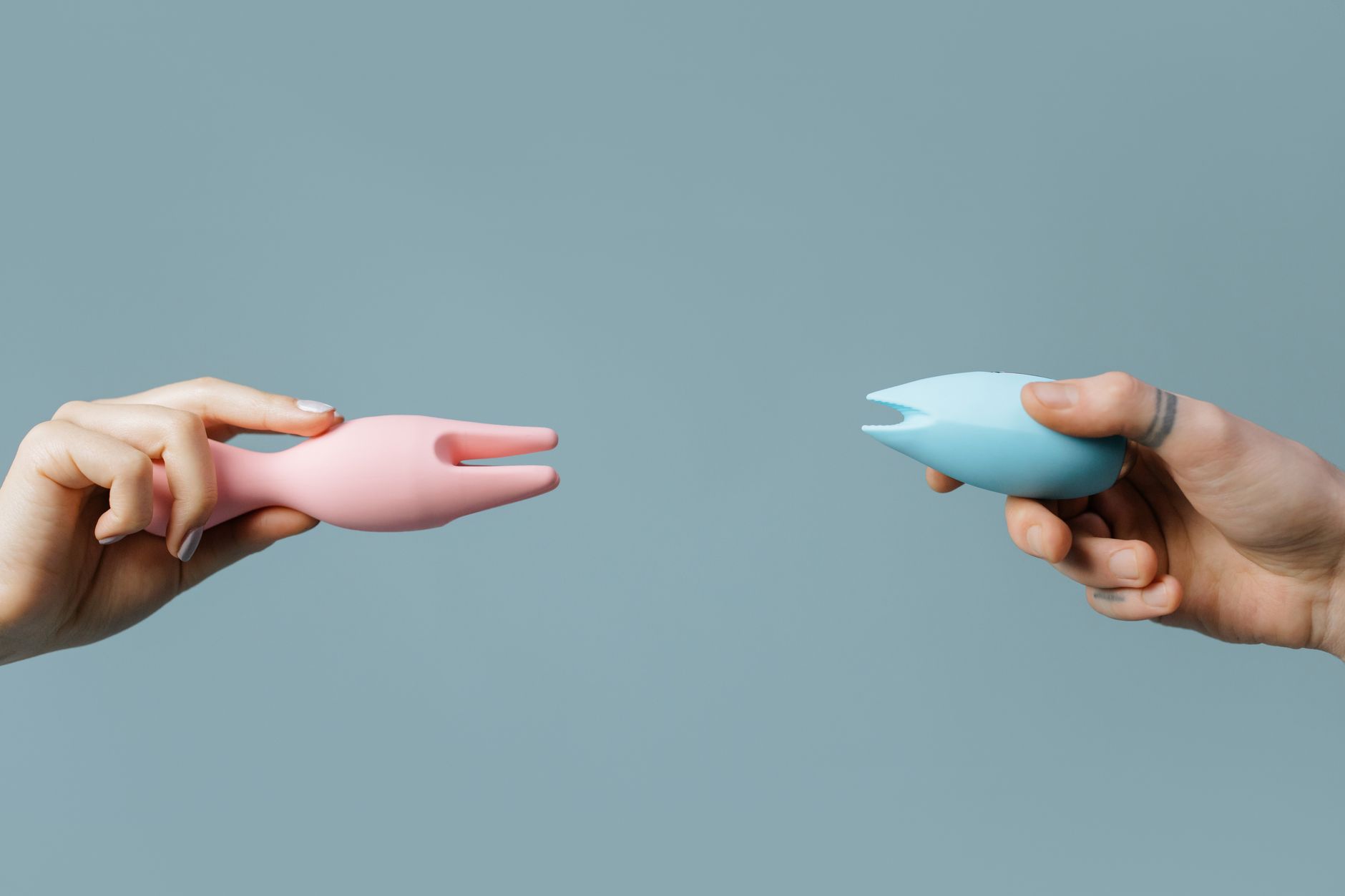 Two hands holding pink and blue clitoral vibrators against a pastel background.