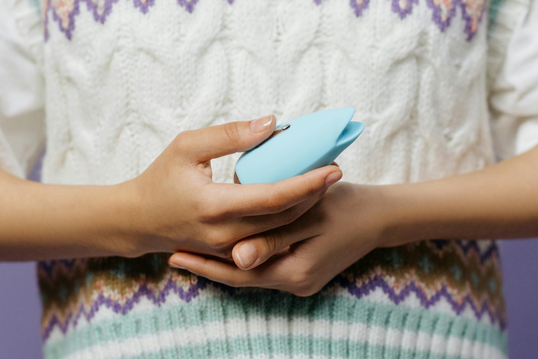 Mãos segurando um vibrador pessoal azul contra um suéter tricotado