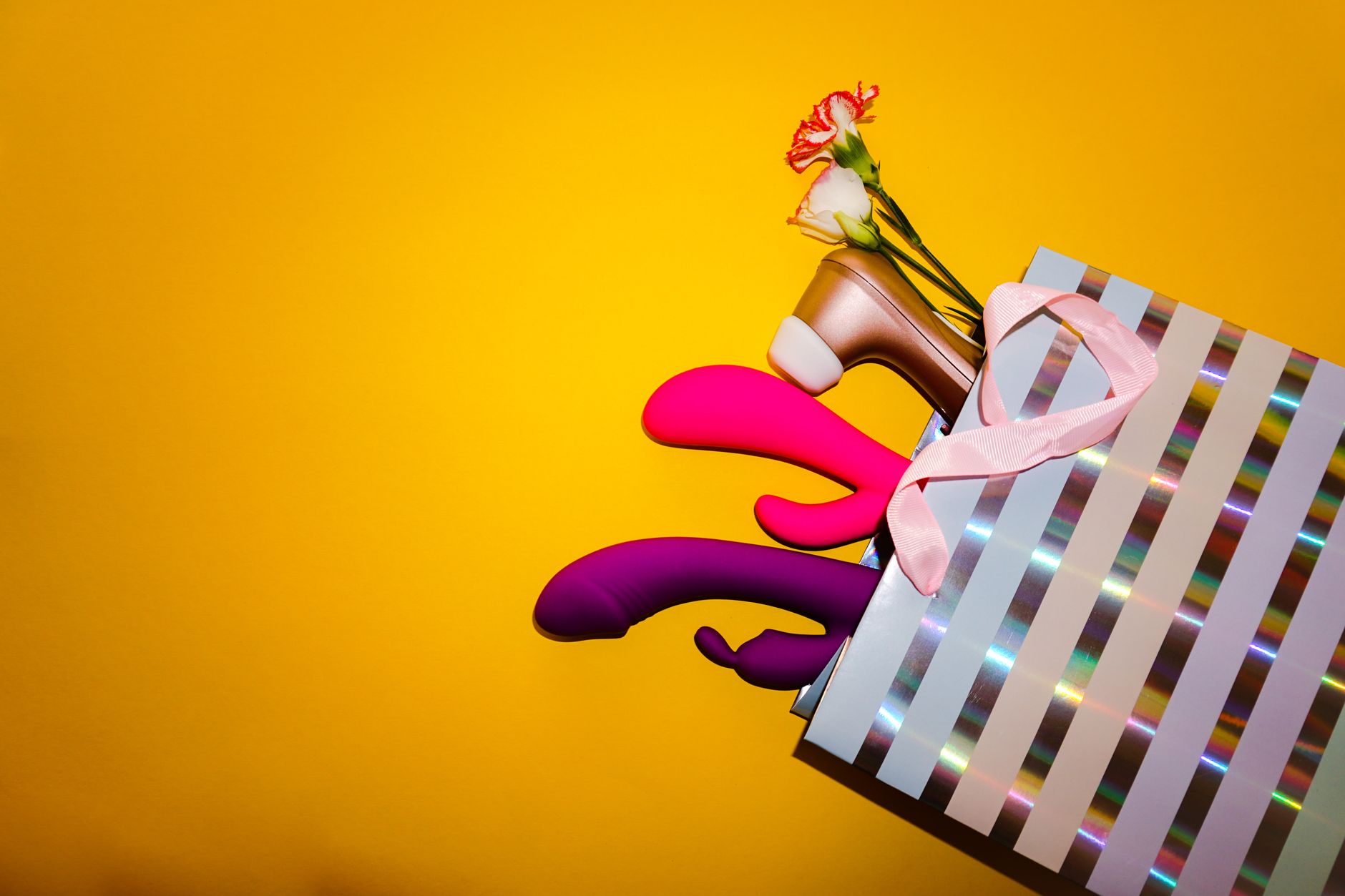 Colorful vibrators with flowers arranged in a holographic gift bag against a bright yellow background.